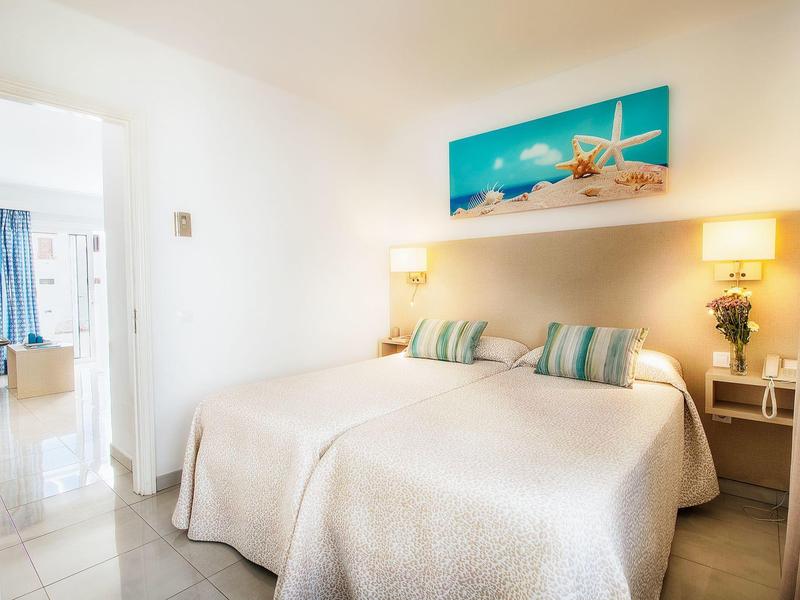 Camera d'hotel luminosa con letto matrimoniale, pareti bianche e quadro della spiaggia sopra la testiera.