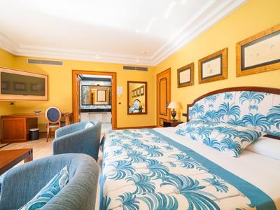 Světly hotelový pokoj s manželskou postelí, modrým posezením a žlutými stěnami zdobenými obrazy.