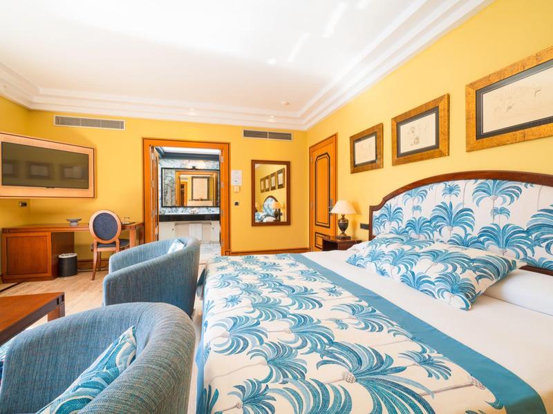 Jasny pokój hotelowy z żółtymi ścianami, łóżkiem w niebieskie wzory i wygodnymi fotelami.