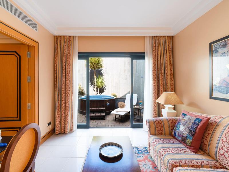 Przytulny pokój hotelowy z sofą, stolikiem i widokiem na taras z palmami.