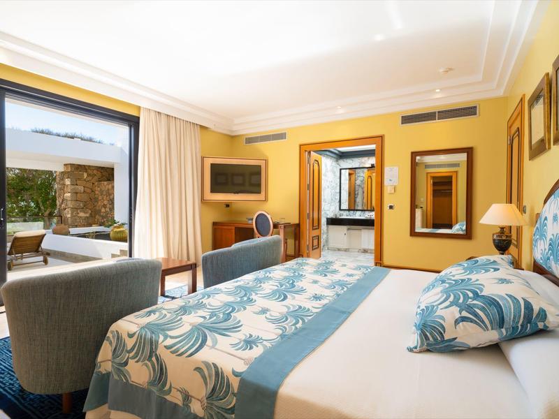 Jasny pokój hotelowy z dużym łóżkiem, fotelami i widokiem na taras.