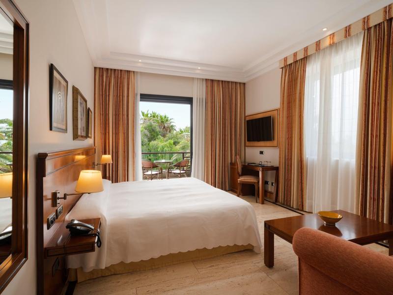 Jasny pokój hotelowy z podwójnym łóżkiem, telewizorem, balkonem i beżowym wystrojem.