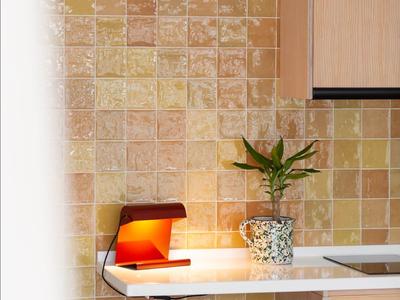 Piccola zona cucina con sgabello, pianta e lampada rossa contro parete piastrellata beige