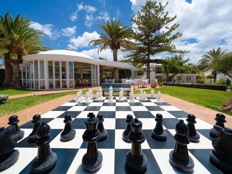 Grande scacchiera all'aperto su una terrazza con palme e edificio moderno sullo sfondo
