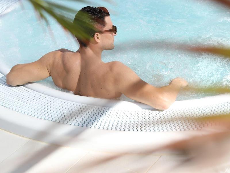 Donna si rilassa in una vasca idromassaggio con occhiali da sole, circondata da foglie di palma.