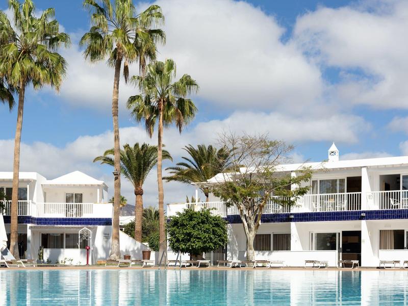 Complesso alberghiero moderno con piscina, palme e ombrelloni sotto un cielo azzurro.