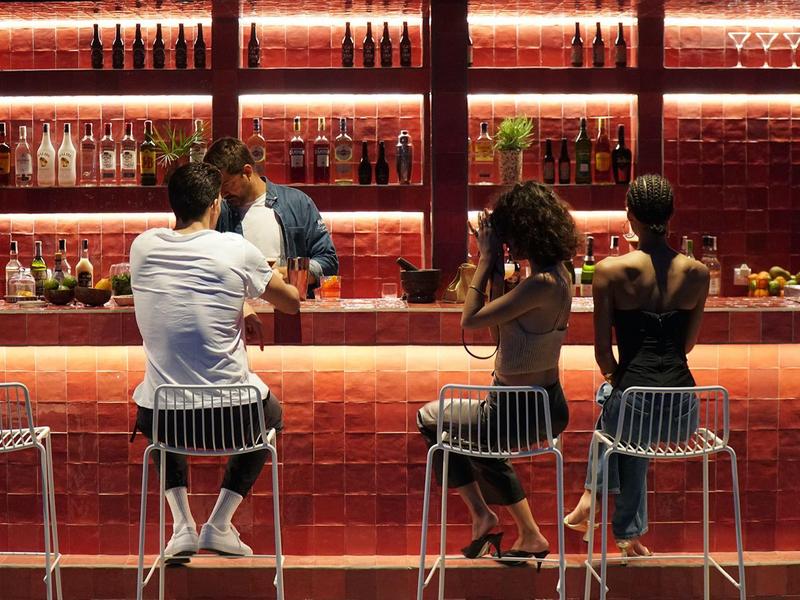 Tre persone sedute al bar con scaffali illuminati di rosso che parlano con il barista.