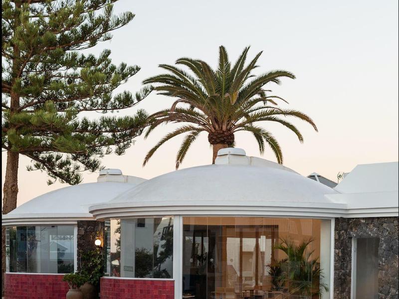 Complesso alberghiero moderno con piscina, palme e grandi finestre di vetro al tramonto.