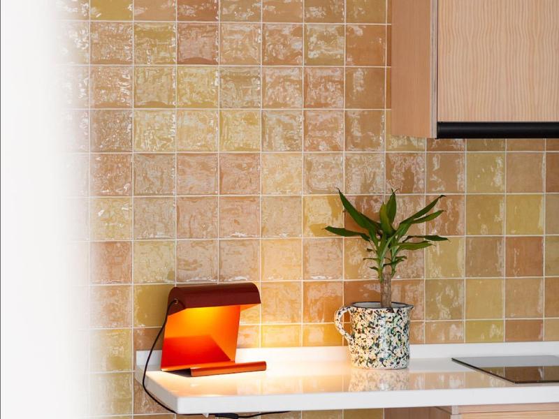 Piccola zona cucina con sgabello, pianta e lampada rossa contro parete piastrellata beige