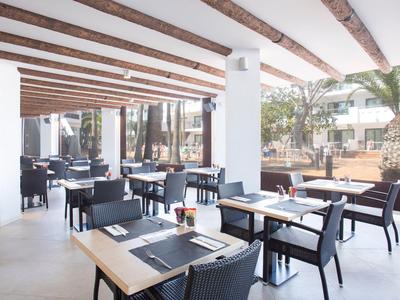 Moderner Außenbereich eines Restaurants mit schwarzen Stühlen, Holztischen und weißen Wänden unter Holzbalken.