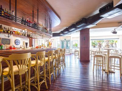 Heller Barbereich mit Holzstühlen, Theke, hohen Tischen und großen Fenstern zum Außenbereich.