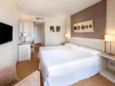 Hell eingerichtetes Hotelzimmer mit zwei weißen Betten, Fernseher an der Wand und Schreibtisch im Hintergrund.