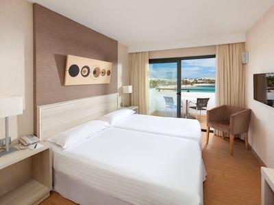 Helles Schlafzimmer mit Doppelbett, Balkon, zwei Sesseln und Blick auf einen See und Hügel.
