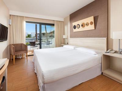 Helles Hotelzimmer mit Doppelbett, Balkon, Fernseher, Sesseln und moderner Holzmöblierung.
