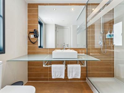 Modernes Badezimmer mit Holzfliesen, Glasablage, weißem Waschbecken und großer Spiegelfläche.