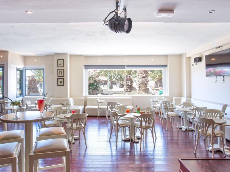 Ristorante luminoso e moderno con pavimento in legno, tavoli e sedie bianchi, ampie finestre.