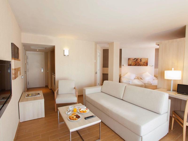 Camera d'hotel luminosa con divano bianco, TV, letto e colazione sul tavolo.