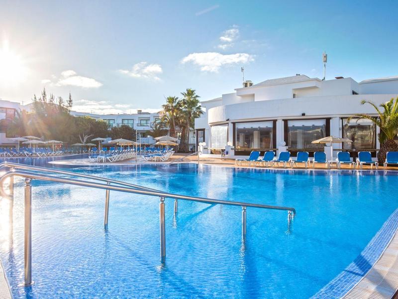 Grande piscina all'aperto con ombrelloni e lettini davanti a un edificio alberghiero bianco in una giornata di sole.
