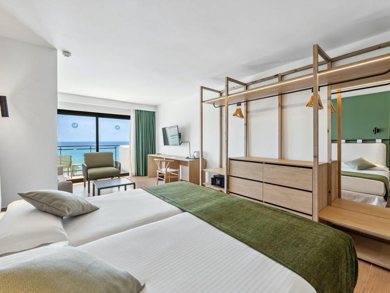 Camera d'hotel luminosa con vista mare, due letti, area salotto e guardaroba aperto.