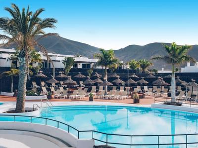 Okrągły basen hotelowy z leżakami i parasolami na tle gór pod czystym niebem.