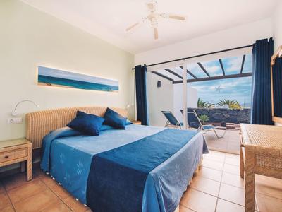 Jasny pokój hotelowy z podwójnym łóżkiem, niebieskimi odcieniami i dostępem do tarasu z leżakami i zielonym widokiem.