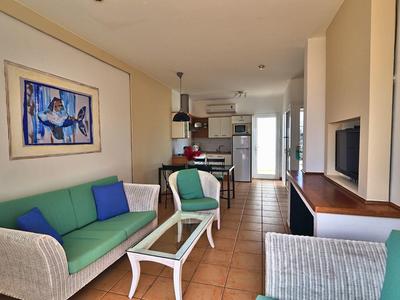 Jasny salon z zielonymi sofami, szklanym stołem i otwartą kuchnią z jadalnią w tle.