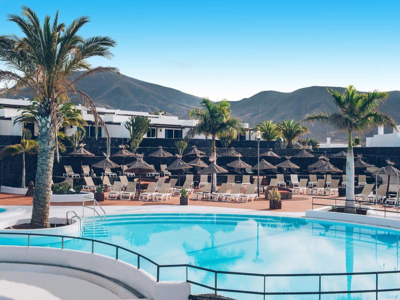 Okrągły basen hotelowy z leżakami i parasolami na tle gór pod czystym niebem.