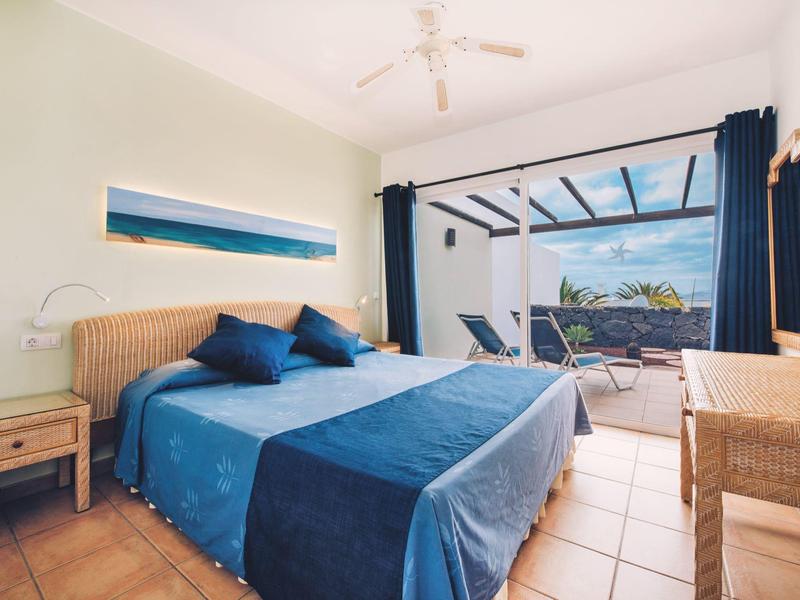 Jasny pokój hotelowy z podwójnym łóżkiem, niebieskimi odcieniami i dostępem do tarasu z leżakami i zielonym widokiem.