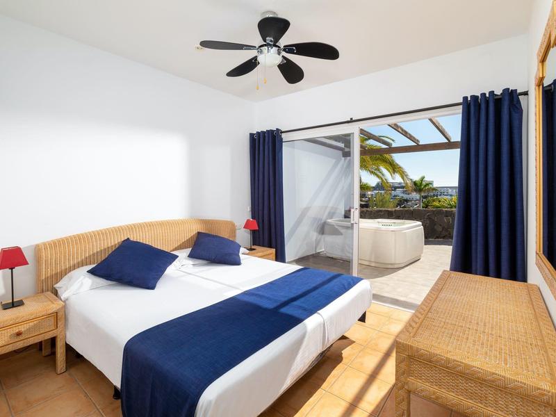 Jasna sypialnia z dużym łóżkiem, niebieskimi akcentami i dostępem do tarasu z jacuzzi.