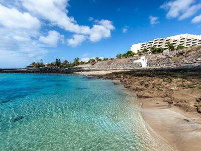 Clear water and rocky beach with hotel building on the shore under blue sky.