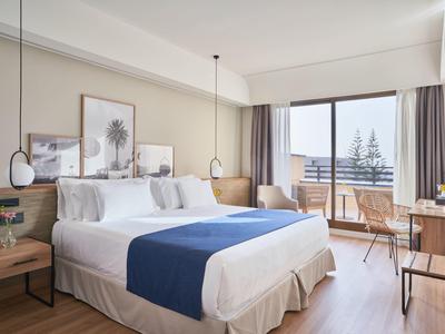 Bright hotel room with large bed, balcony, and sea view in daylight.