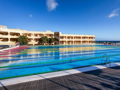 Großer Außenpool mit Schwimmbahnen vor einem Hotelgebäude unter blauem Himmel.