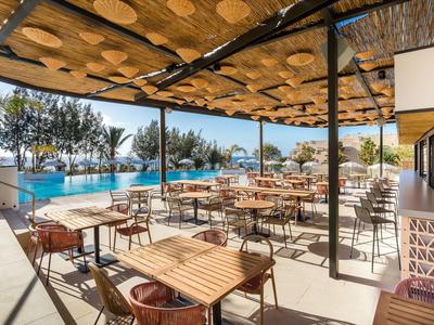 Covered terrace with wooden tables and chairs beside a pool with palm trees in the background.
