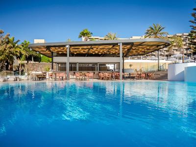 Klare blaue Poollandschaft vor einer offenen Bar mit Sonnendach und Palmen.