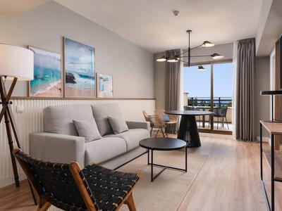 Modern living room with sofa, armchair, dining table, and balcony overlooking the sea.