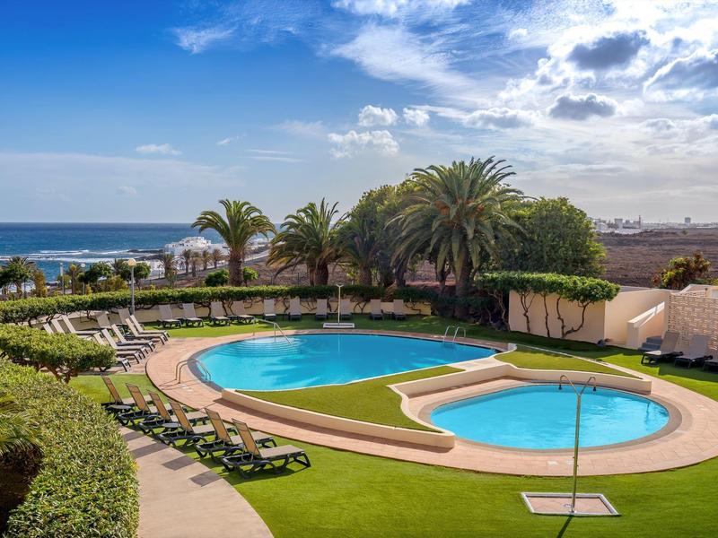 Tranquil hotel pool with lounge chairs, palm trees, and ocean view under a blue sky.