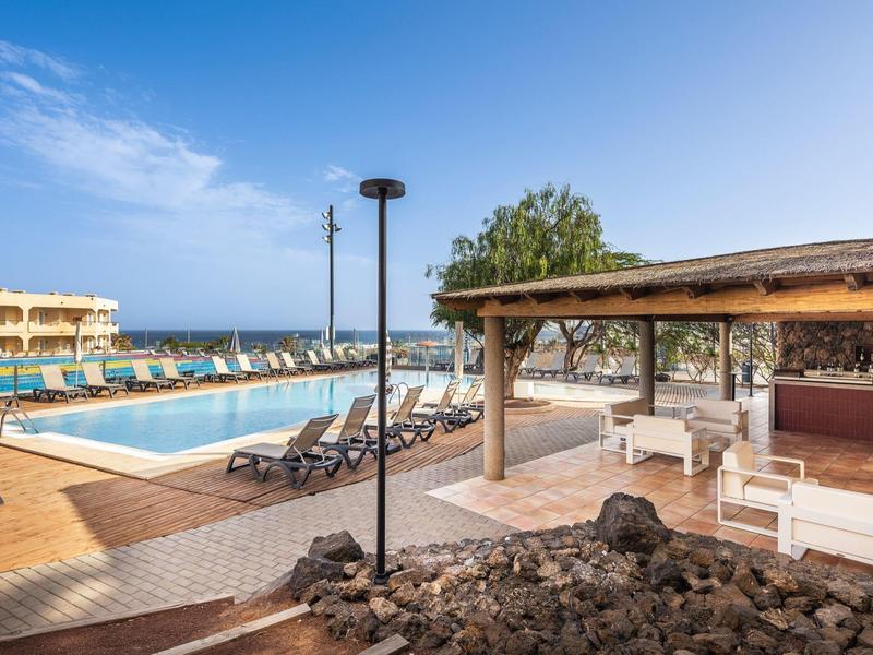 Hotel with pool, sun loungers, and bar area under clear blue sky by the sea.