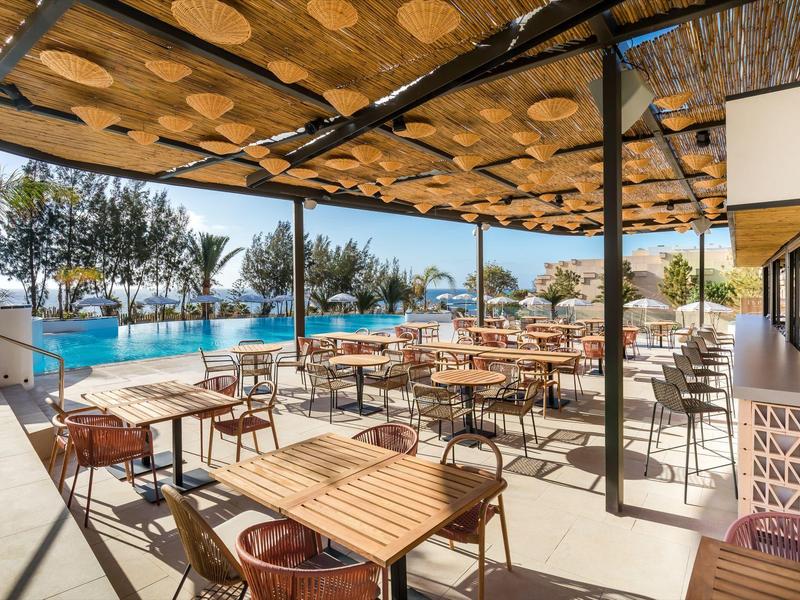 Covered terrace with wooden tables and chairs beside a pool with palm trees in the background.