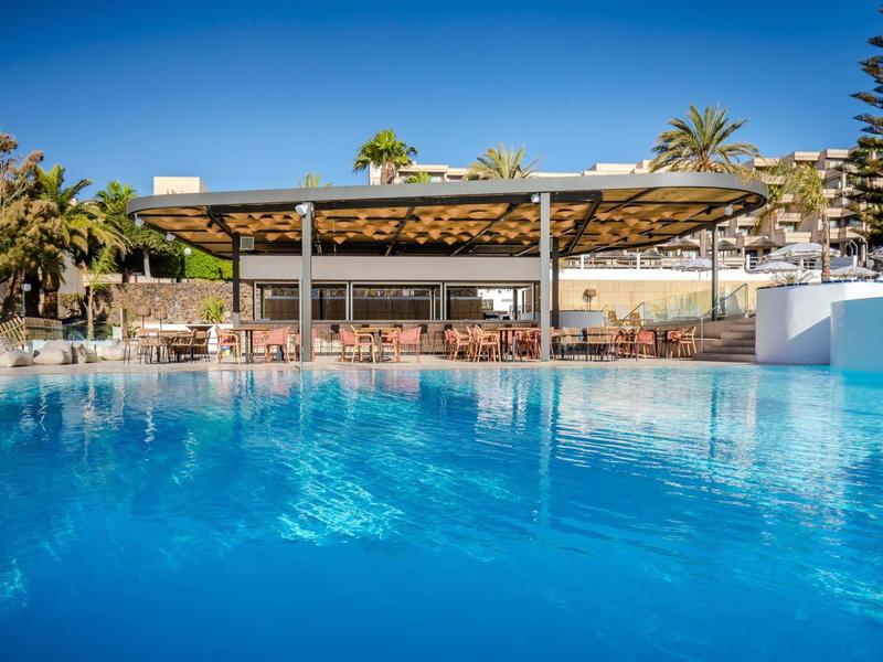 Large pool with clear water in front of a covered pool bar and palm trees in the background.