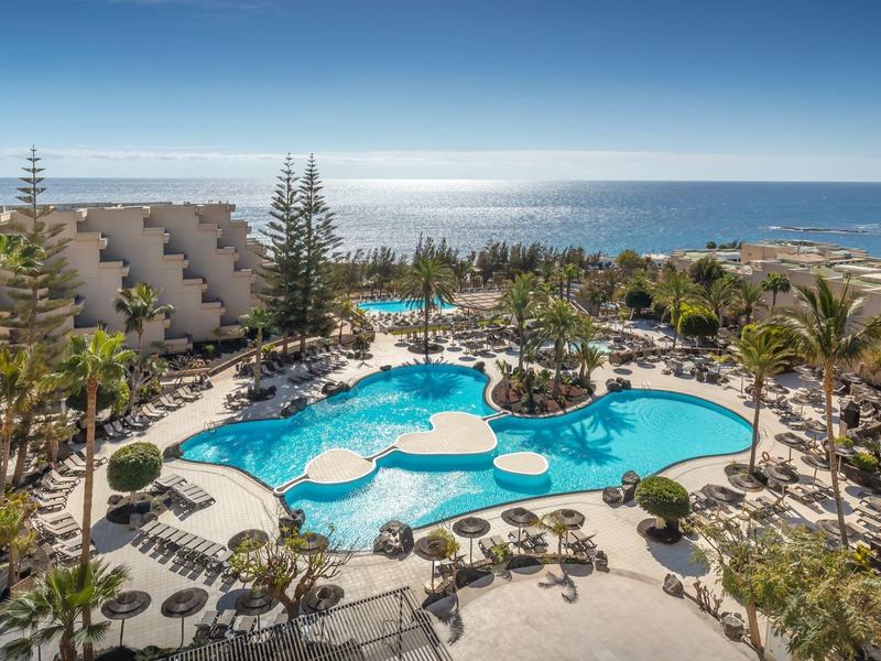 Large hotel pool with sun umbrellas, palm trees, and ocean view in the background.