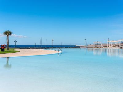 Großer, klarer Pool mit Palmen und Blick aufs ruhige, blaue Meer unter strahlend blauem Himmel.