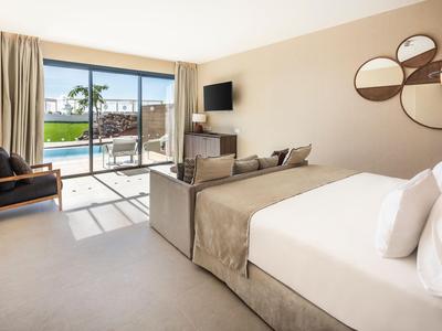 Modernes Hotelzimmer mit großem Bett, Sitzecke und Blick auf Pool und Palmen durch eine Glas-Schiebetür.