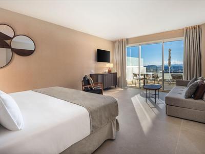 Helles Hotelzimmer mit großem Bett, Sofa, TV und Balkon mit Blick auf das Meer.