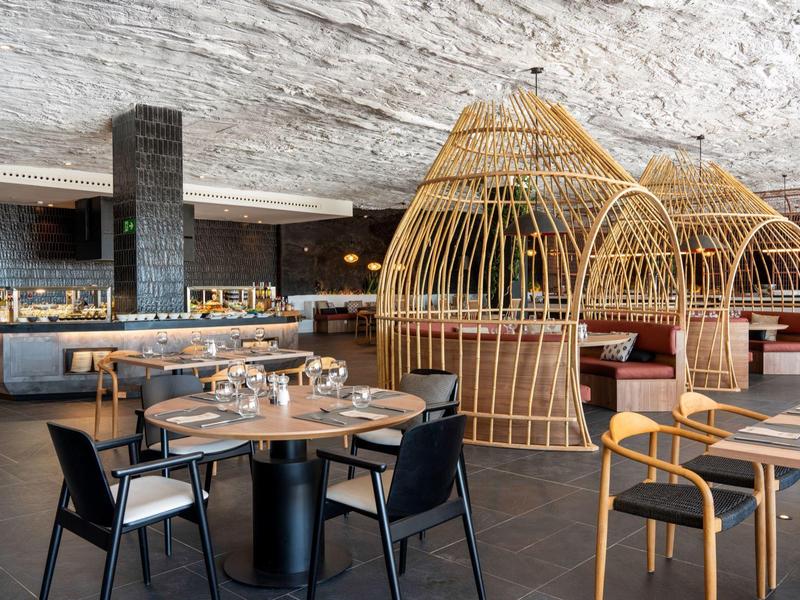 Nowoczesna restauracja z okrągłymi drewnianymi stołami, czarnymi i naturalnymi krzesłami oraz plecionymi pawilonami.