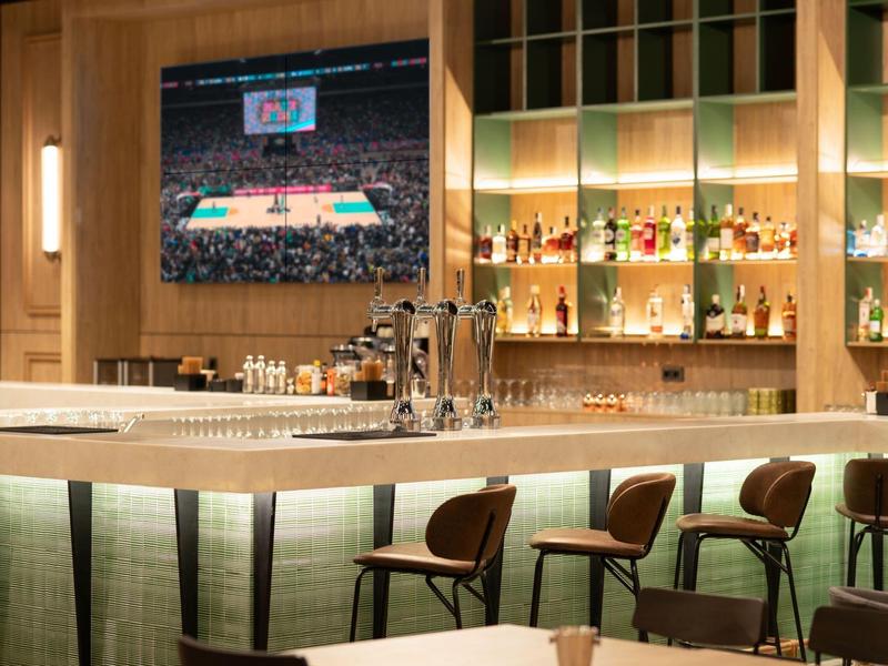 Nowoczesny bar hotelowy z jasnym barem, wygodnymi stołkami barowymi i telewizorem na ścianie.