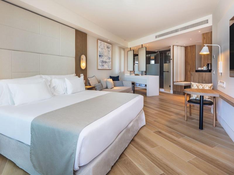 Nowoczesny pokój hotelowy z dużym łóżkiem, częścią wypoczynkową i drewnianą podłogą w neutralnych tonach.