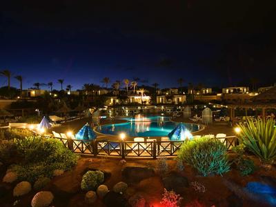 Vista notturna di una piscina illuminata in un complesso alberghiero con piante e palme.