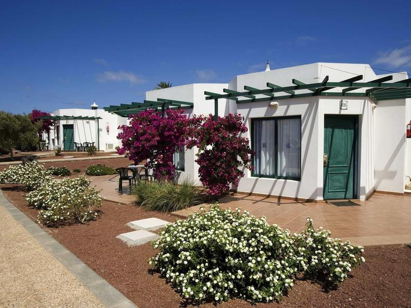Bungalows blancs avec portes vertes et buissons en fleurs sous un ciel ensoleillé.