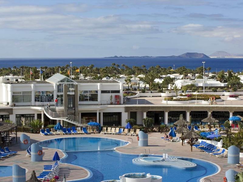 Grande piscine d'hôtel avec chaises longues bleues et vue sur la mer sous un ciel nuageux.