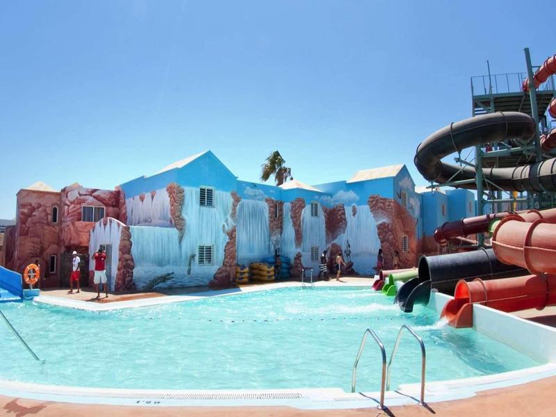 Parc aquatique avec mur de cascade, toboggans et piscine sous un ciel bleu clair.